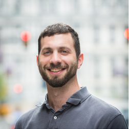 A man with short dark hair and a beard, wearing a gray polo shirt, stands outdoors smiling with a blurred city background.