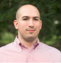 A person with a shaved head, wearing a light pink button-down shirt, stands outdoors in front of blurred greenery.