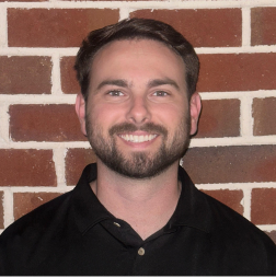A man with short brown hair and a beard smiles at the camera, standing in front of a brick wall and wearing a black collared shirt.