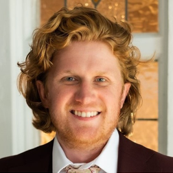 A man with wavy red hair and a beard, wearing a maroon suit and patterned tie, smiles in front of a window with frosted glass panes.