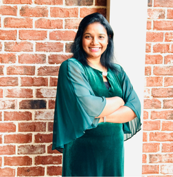 Woman in a green dress stands with arms crossed, smiling, in front of a red brick wall.