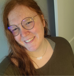 Person with long reddish hair and glasses smiles at the camera while wearing a dark green textured shirt and a necklace with a molecular structure pendant.
