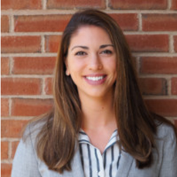 A woman with long brown hair, wearing a gray blazer and striped blouse, stands smiling in front of a red brick wall.