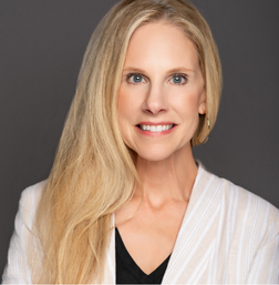 A woman with long blonde hair, wearing a white blazer over a black top, smiles at the camera against a plain gray background.