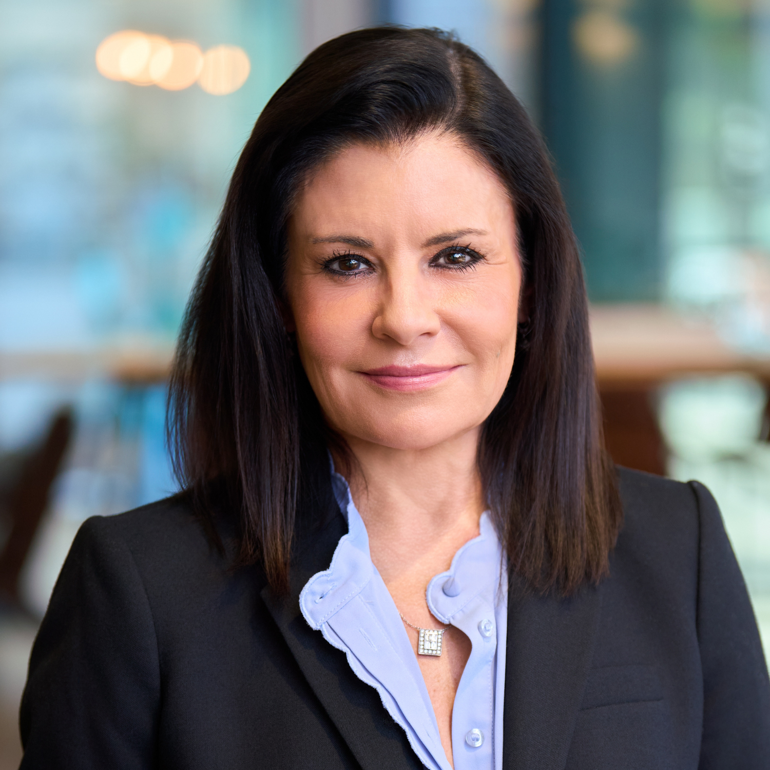 A woman with straight dark hair wearing a black blazer and light blue blouse is standing indoors, looking at the camera with a neutral expression.
