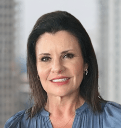 Woman with dark hair and a blue blouse smiling, standing in front of a blurred cityscape background.