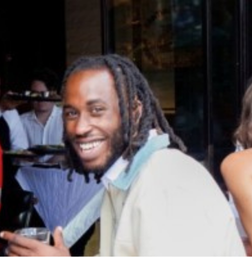 A man with dreadlocks smiles at the camera while sitting at a table in an indoor restaurant setting.