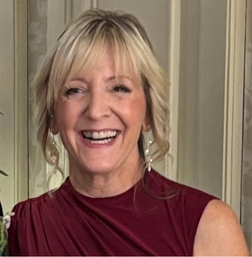 A smiling woman with blonde hair wearing a sleeveless burgundy dress stands indoors in front of a light-colored patterned wall.