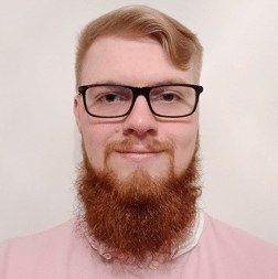 A young man with a red beard and glasses is facing the camera and smiling. He is wearing a light pink shirt and has short, light hair. The background is plain white.