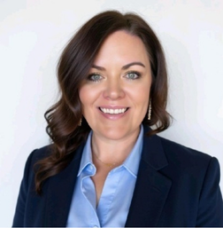 Woman with long brown hair wearing a navy blazer and light blue blouse, smiling in front of a plain white background.