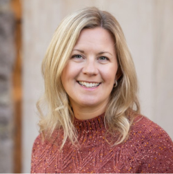 A woman with shoulder-length blonde hair, wearing a textured rust-colored sweater, smiles at the camera against a neutral background.
