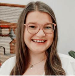 A woman with long brown hair and pink glasses smiles at the camera. She is wearing a white top and a necklace, with a woven wall hanging in the background.