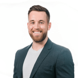 A man with a beard and short hair is smiling, wearing a dark blazer over a light shirt, standing in front of a plain white background.