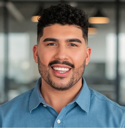 Smiling man with short curly hair and a trimmed beard, wearing a blue button-up shirt, standing in an office setting with blurred background.