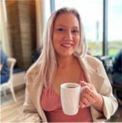 A woman with long light hair sits indoors by a window, smiling and holding a white mug. She wears a light jacket over a pink top.