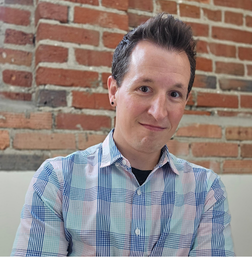 Person with short dark hair wearing a pastel plaid shirt stands in front of a red brick wall, looking at the camera with a slight smile.