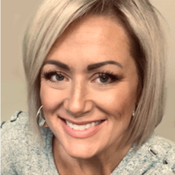 Woman with short blonde hair and light makeup, smiling at the camera while wearing a light-colored top.