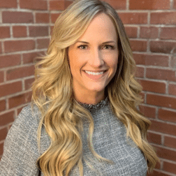 A woman with long blonde hair and a grey top smiles in front of a brick wall.