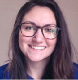Woman with long brown hair and glasses smiling at the camera, wearing a blue top, with a neutral background.