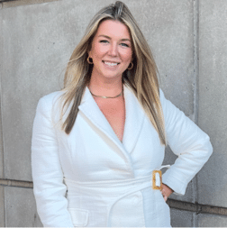 A woman with long blonde hair wearing a white blazer stands in front of a concrete wall, smiling at the camera.