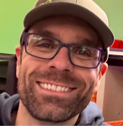 A man wearing glasses and a cap smiles at the camera.