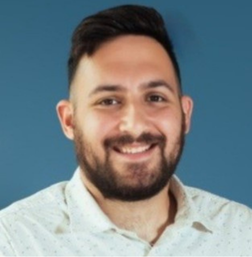 A man with dark hair and a beard, smiling, wearing a white collared shirt, poses in front of a plain blue background.