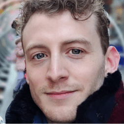 A person with short curly hair and blue eyes is looking at the camera, wearing a dark coat, with a blurred outdoor background.