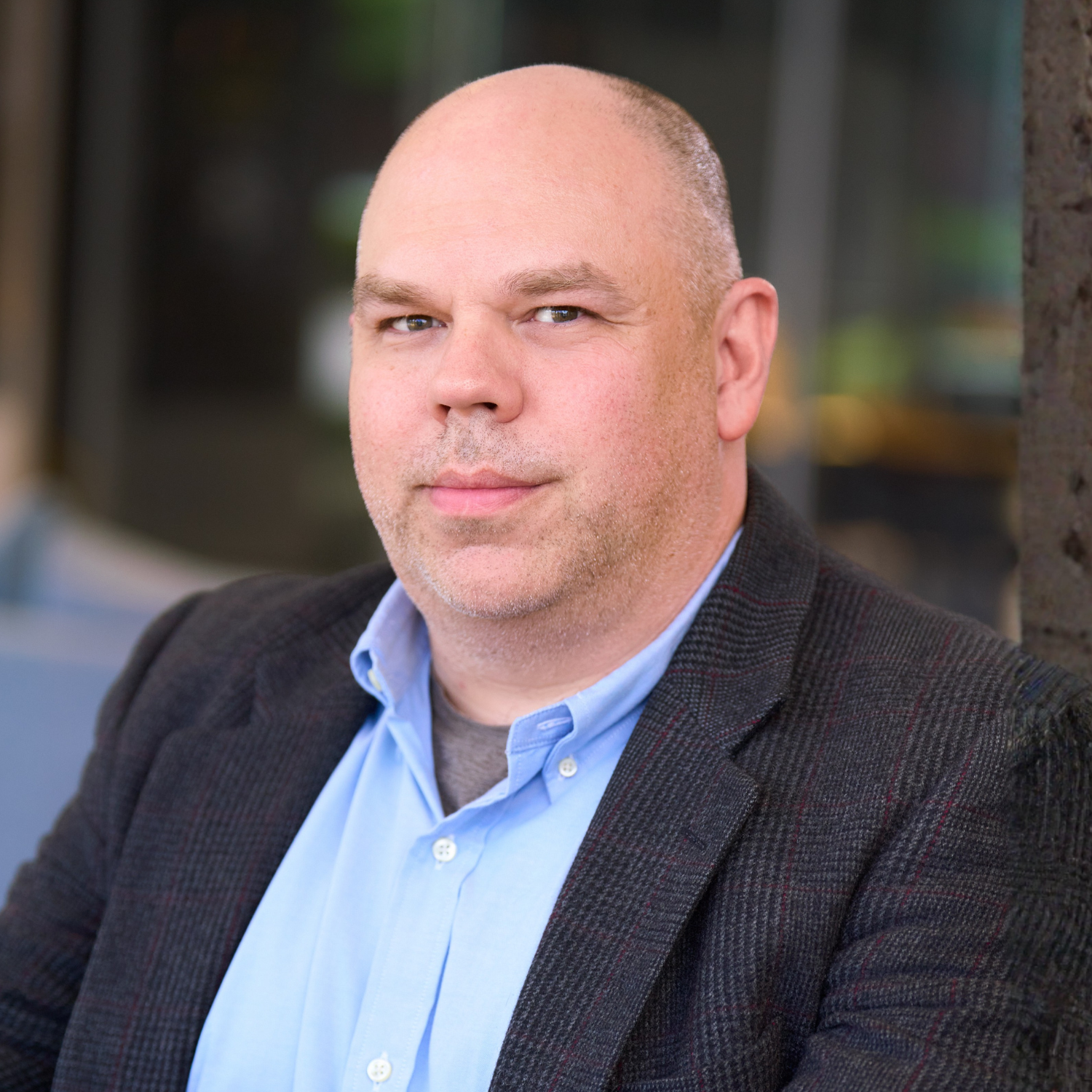 A man with a shaved head in a blue shirt and dark blazer sits indoors, looking at the camera with a neutral expression.