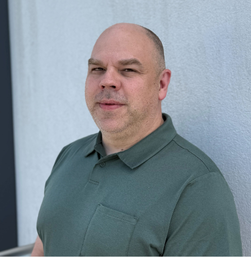 A man with a shaved head and stubble, wearing a green polo shirt, stands in front of a light-colored textured wall, looking slightly past the camera.