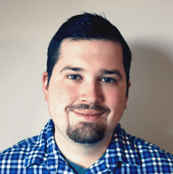 Man with short dark hair and a trimmed beard, wearing a blue plaid shirt, smiling slightly, against a plain light-colored background.