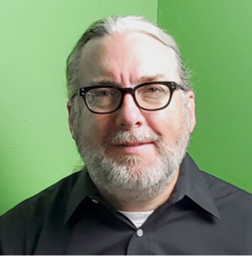 A middle-aged person with gray hair, beard, and glasses, wearing a black shirt, is standing in front of a green wall and smiling slightly.
