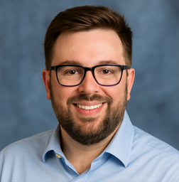 Man with short brown hair, beard, and glasses, wearing a light blue collared shirt, smiling in front of a blue background.