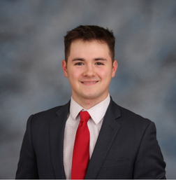 A young man in a dark suit, white shirt, and red tie is smiling in front of a gray and blue mottled background.