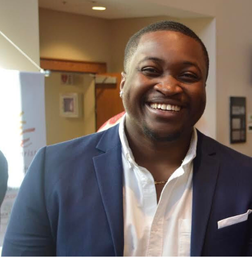 A man in a navy blazer and white shirt smiles while standing indoors at an event or conference.