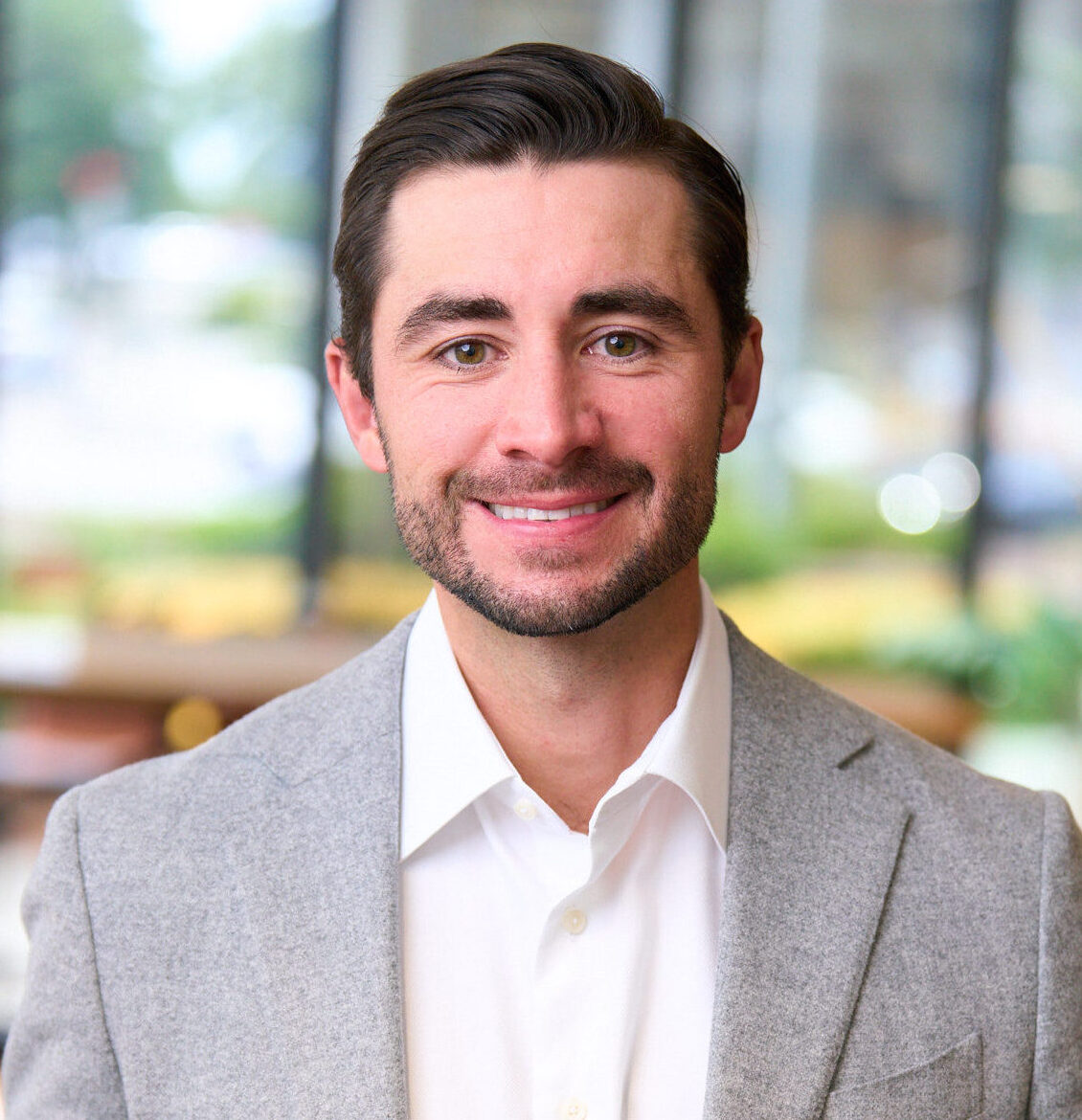 A man with short brown hair and a trimmed beard, wearing a light gray blazer and white shirt, smiling in a brightly lit indoor setting.