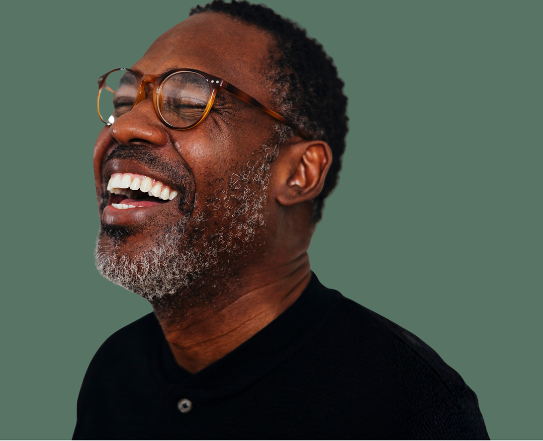 A middle-aged man with glasses and a beard laughs, looking to the side, against a plain green background.