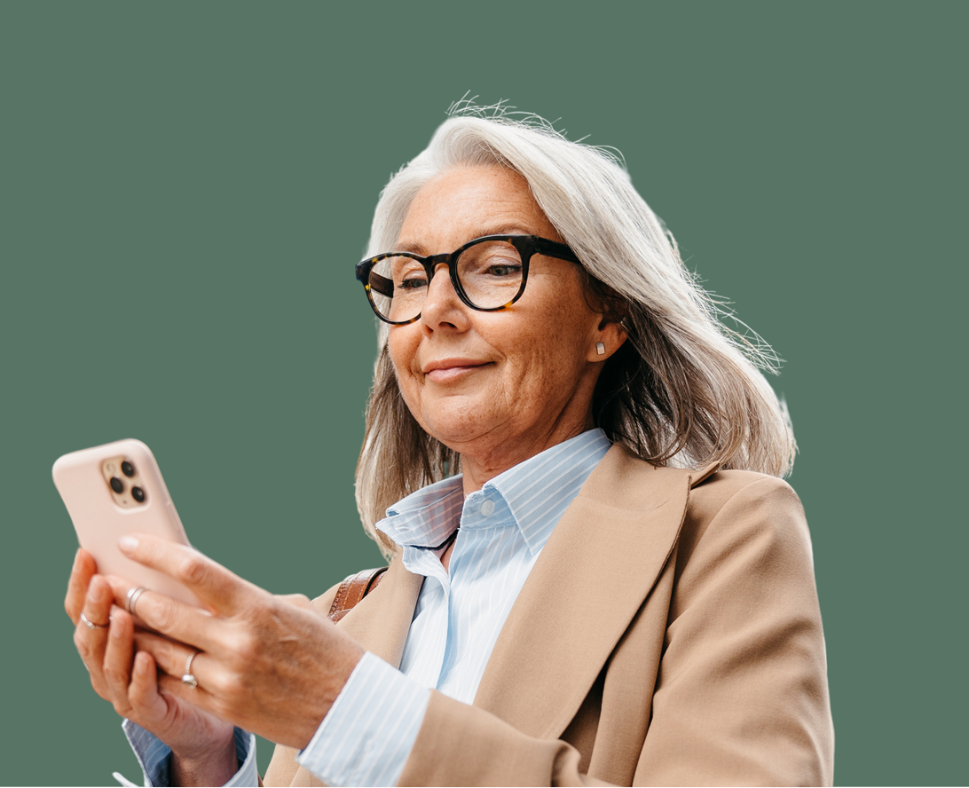 Older woman with gray hair and glasses, wearing a tan blazer and blue shirt, looks at a smartphone against a plain green background.