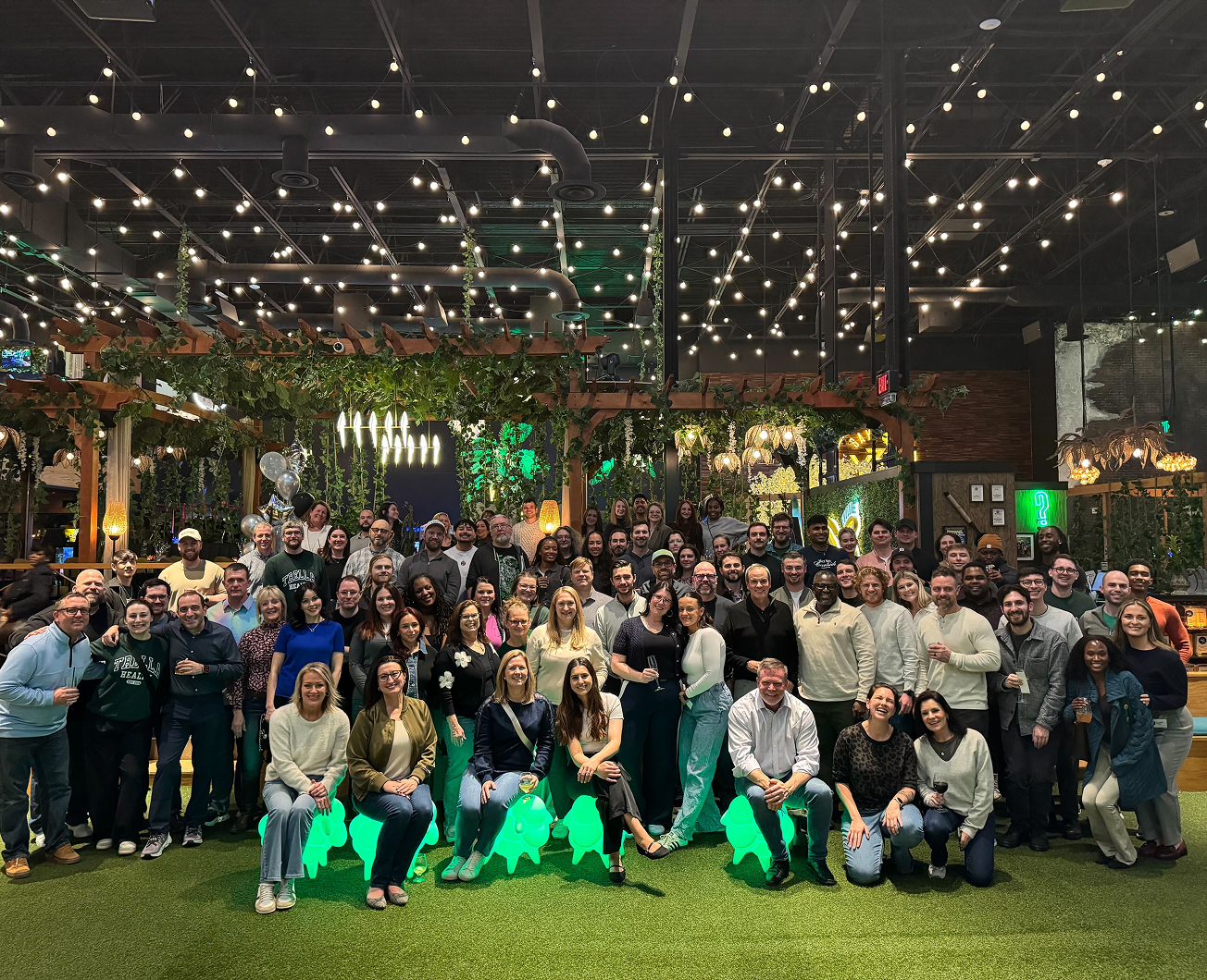 Large group of people posing together indoors under string lights, with greenery and decorative lighting in the background.