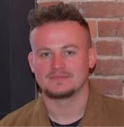 A person with short curly hair and a trimmed beard is facing the camera, standing in front of a brick wall.