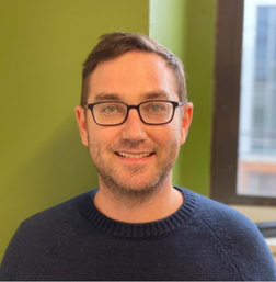 A man with short brown hair, glasses, and a navy sweater smiles in front of a green wall and window.