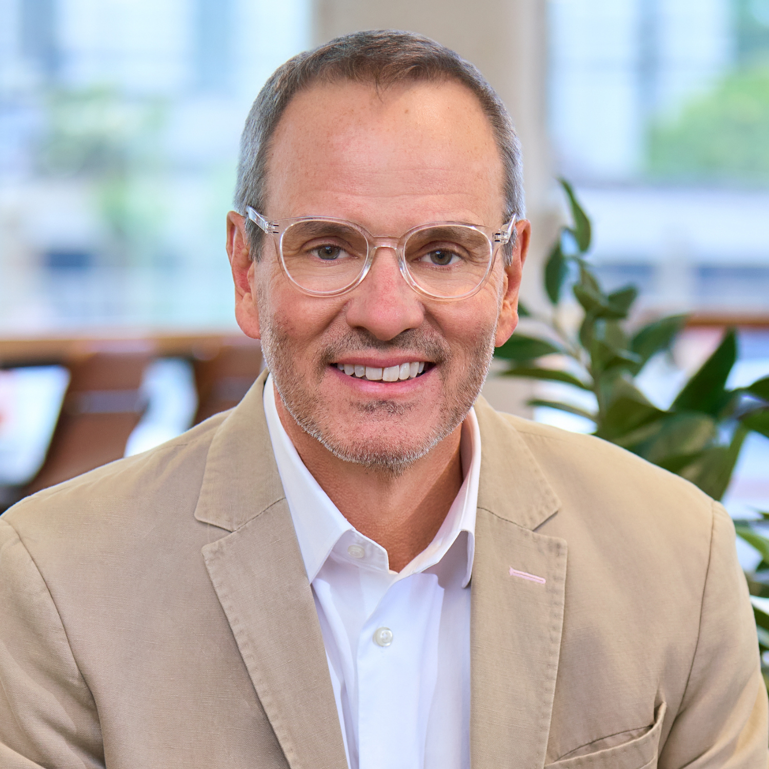 A middle-aged man with short hair and glasses, wearing a light beige blazer and white shirt, smiles in an office setting with blurred windows and plants in the background.