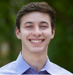 A young man with short brown hair, wearing a light blue collared shirt, smiles at the camera with a blurred green background.