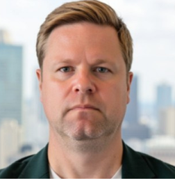 A man with short light brown hair and a serious expression stands in front of a blurred cityscape background.