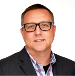 Man with short brown hair and glasses wearing a suit jacket over a plaid shirt, facing forward against a plain white background.