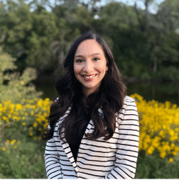 A woman with long dark hair, wearing a white and black striped blazer, stands outdoors in front of yellow flowers and green trees.
