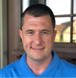 A man with short brown hair wearing a blue collared shirt is smiling in front of a wooden window.