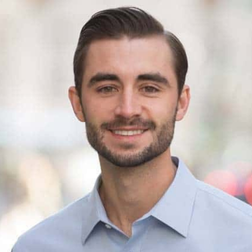 A man with short dark hair and a beard, wearing a light blue collared shirt, smiles at the camera against a blurred outdoor background.
