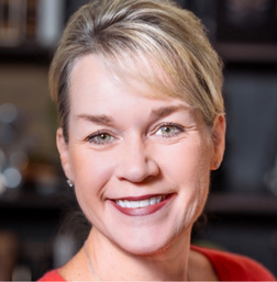 A woman with blond hair pulled back smiles at the camera, wearing a red top. The background is softly blurred.