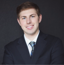 Young man in a dark pinstripe suit and striped tie poses against a plain dark background, smiling with arms crossed.