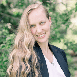 A woman with long, wavy blonde hair wearing a dark blazer and white top smiles outdoors in front of green foliage.